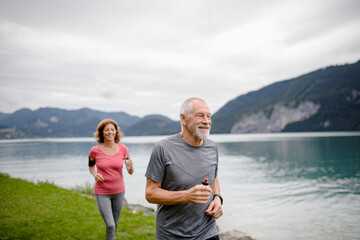 Senior couple running by lake in nature. Elderly husband and wife spending active vacation in the mountains, enjoying physical activity and relaxation outdoors.