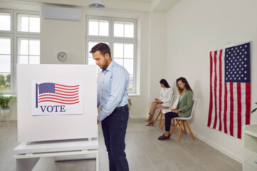 Side view portrait of young american voter man standing at vote center with USA flags in voting...