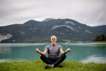 Naklejka na ściany i meble Senior man doing outdoor yoga, meditating by the lake in mountains. Elderly sportsman spending active vacation in nature.