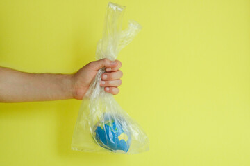 a man's hand holds the globe in a plastic transparent bag on a yellow background, abstraction on the theme of ecology for Earth Day, environmental problems, pollution of the planet with plastic bags