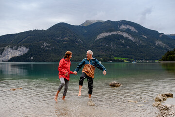 Active elderly couple hiking together in spring mountains, walking in cold water in mountain lake....