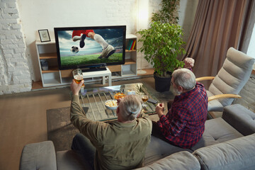 Elderly men, sport fans, friends sitting in living room at hoe, drinking beer and watching online...