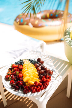 Delicious juicy fruit and berry set on the table by the pool. Pineapple, cherries, strawberries, raspberries and grapes on a plate next to the chaise longue. Tasty snacks for lunch.