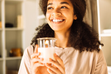 Close-up of charming mixed-race teenager holding burn candle in glass candlestick and laughing....