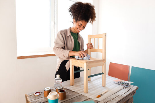 A beautiful smiling mixed-race girl wipes a wooden chair with a rag.