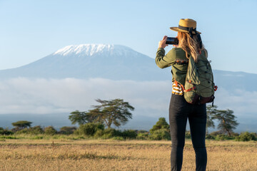 beautiful young girl In a hat stands against the backdrop of the Kilimanjaro volcano and looks away. The concept of tourism and African safari. A female backpacker takes pictures of nature and animals