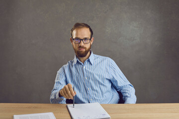 Portrait of a confident man working with paper documents or preparing for an exam. Bearded middle-aged businessman with a pencil in his hands sitting at a table on a gray concrete background.
