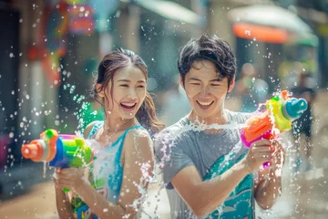 Selbstklebende Fototapeten Happy traveler asian man and woman wearing summer shirt holding colourful squirt water gun over blur city, Water festival holiday concept © grapestock