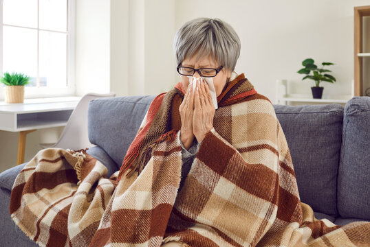 Sick with flu. Unhappy frozen mature woman sick at home and suffering from rhinitis, cold and influenza. Caucasian senior woman is sitting on bed wrapped in plaid and wiping her nose with paper napkin