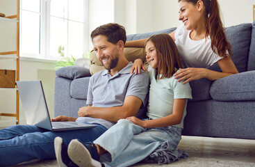 Happy young family of three with child girl sitting on the floor at home using laptop together having video call talking online. Smiling parents with daughter watching funny cartoon or movie.