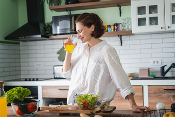 Young caucasian smiling woman drinking orange juice while standing in the kitchen at home, vitamins and healthy eating - 779522316