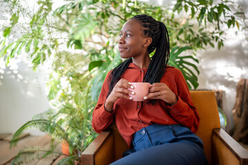 Relaxed dreaming african american woman holding cup of coffee sitting on chair in urban jungle...