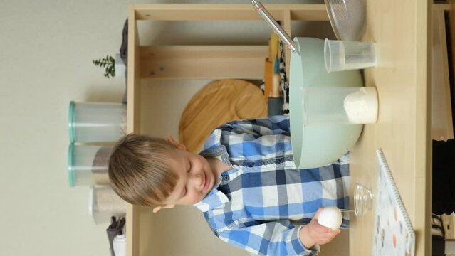 A two-year-old boy cracks a raw egg into dough. Toddlers in the kitchen, vertical slow motion.