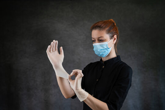 Woman Doctor Or Nurse In Black Uniform And Protective Mask Putting Protective Glove Isolatedon Dark