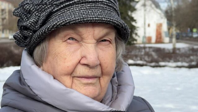 Portrait of serious mature old woman dressed in a grey coat and black hat in the background of the park in winter season. The woman looks away