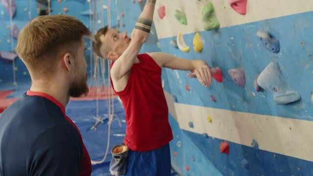 Young male athlete with one leg rubbing hands with chalk and climbing wall with friend while training together in indoor gym