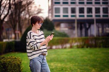 Engrossed in her phone, a young woman in a striped sweater stands in a vibrant city park, her style...