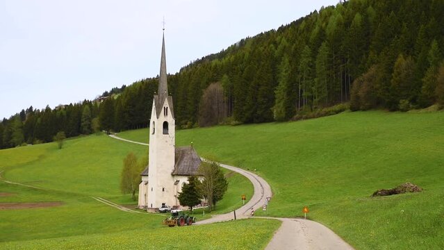 Spring rural landscape with church and tractor driving to agricultural field in Villabassa village in Dolomites Alps, South Tyrol, Italy. Chiesa Madre di Santa Maria Maddalena in alpine meadow