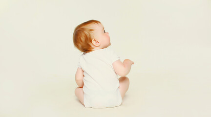 Happy cute baby sitting on the floor on white studio background, looking up, back view