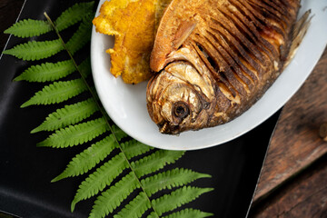 Closeup shot of a plate with a whole fried fish and plantains on a board with fern leaves © Wirestock
