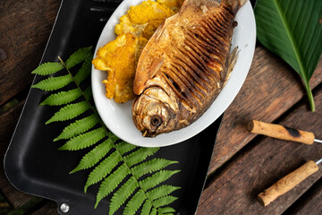Top view of a whole cooked fish and fried plantains on a plate on a black board with a fern branch