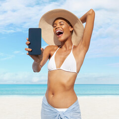 Happy young woman at the beach side showing mobile phone, wearing a turquoise sun hat and bikini, portrait of African latin American woman in sunny summer day with blue sky, concept of summer holiday - obrazy, fototapety, plakaty