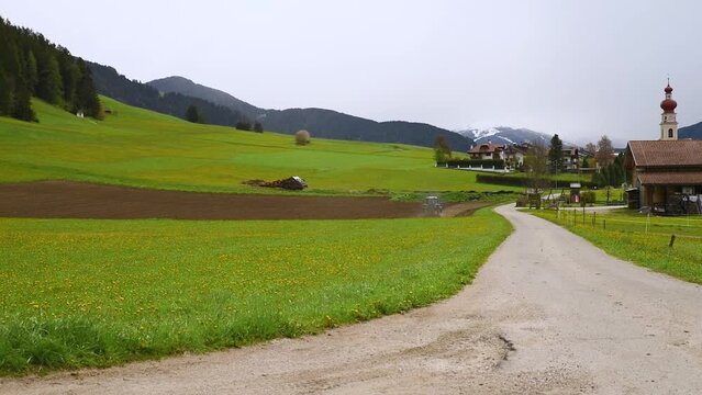 Spring rural landscape with working tractor on agricultural field, road and church in Villabassa village in Dolomites Alps, South Tyrol, Italy