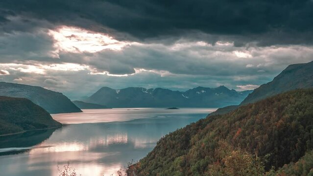 Rays of the setting sun shine through stormy clouds over a calm fjord in a time-lapse video.