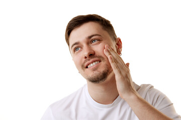 Young handsome man in white t-shirt looking away while applying facial-care serum against white studio background. Concept of natural beauty, male health, anti-aging, spa procedures, cosmetic product.