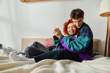 cheerful handsome man with headphones hugging his girlfriend lovingly while at home on bed