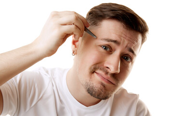 Young man in white T-shirt plucks his eyebrows with tweezers while looking at camera against white studio background. Concept of beauty, male health, anti-aging, spa procedures, cosmetic product