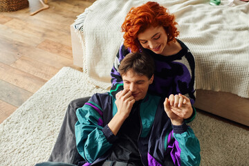 passionate red haired woman in cozy attire smiling and hugging her loving handsome boyfriend - obrazy, fototapety, plakaty