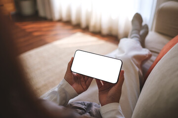 Mockup image of a woman holding mobile phone with blank desktop white screen while sitting on a sofa at home