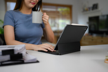 Young woman in casual clothes drinking coffee working online on digital tablet at home