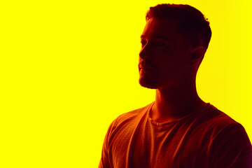 Silhouetted man in T-shirt posing in red neon light against vibrant yellow studio background. Concept of natural beauty, male health, anti-aging, spa procedures, cosmetic products.