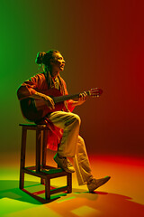 Talented young man, musician with dreadlock sitting on chair and playing guitar against gradient...