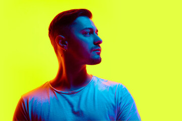 Serious young athletic man posing in casual attire in neon light against vibrant yellow studio...