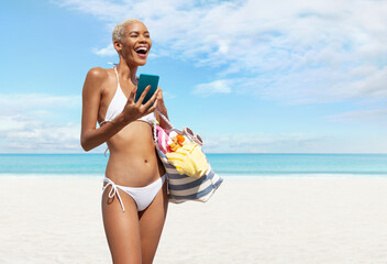 Happy woman at the beach side wearing bikini holding a beach bag and using mobile phone in a sunny day with blue sky. Concept of summer beach holiday, shopping online, booking travel and resort