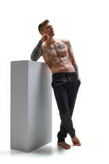 Barefoot, tattooed young man leaning on gray block posing deep in thought against white studio...