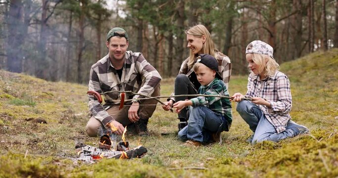 family with children grilling sausages on wooden skewers over a bonfire while camping in forest. nature adventure, bonding activities