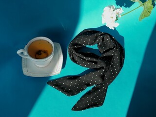 A composition of a cup of herbal tea with a saucer and a silk dark blue polka dot neckerchief and a white flower on a bright blue background with rays of sunlight and shadows     