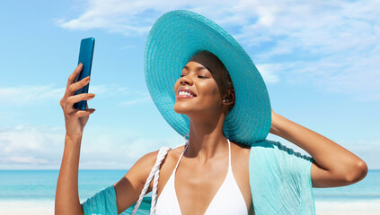 Happy woman at the beach side wearing bikini, blue sun hat and pareo and using mobile phone in a sunny day with blue sky.Concept of summer beach holiday, shopping online, booking travel and resort