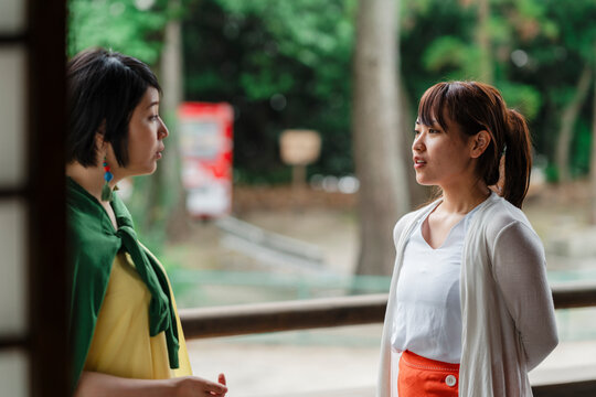 An intense moment as two Japanese women engage in a serious discussion on an urban balcony, with a cityscape in the background