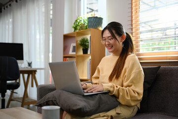 Young woman in casual clothes using laptop sitting on sofa at cozy home