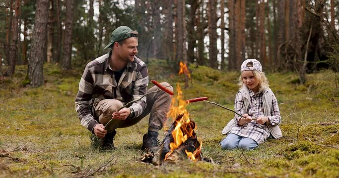 father and daughter together frying sausages over a bonfire while camping in forest. nature adventure, bonding activities