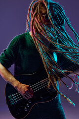 Portrait of stylish guy with dreadlocks, musician playing guitar against dark purple background in...