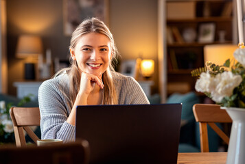 Smiling young woman working at home using laptop while sitting in her living room. - obrazy, fototapety, plakaty
