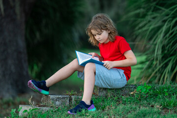 Cute little boy reading book in park. Kid sit on grass and reading book. Kid boy reading interest...