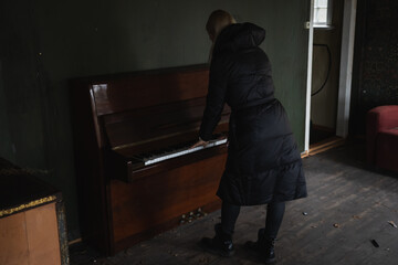 A girl in a black coat plays an old piano in an abandoned house.