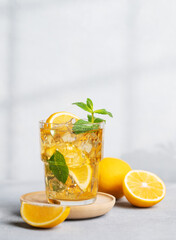 Iced drink with lemon, mint in a glass on a on a light background with shadow and citrus fruits. The concept of a cold tea or lemonade on a hot summer day. Front view.
